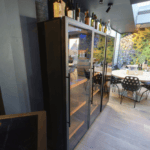 Interieur van een stijlvol café met een bloemenmuurontwerp, glazen deuren, zwarte stoelen en gevlekte tafelbladen bij een bar.