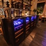 Interior of a dimly lit restaurant with blue light wine coolers, wooden shelves and intimate seating.