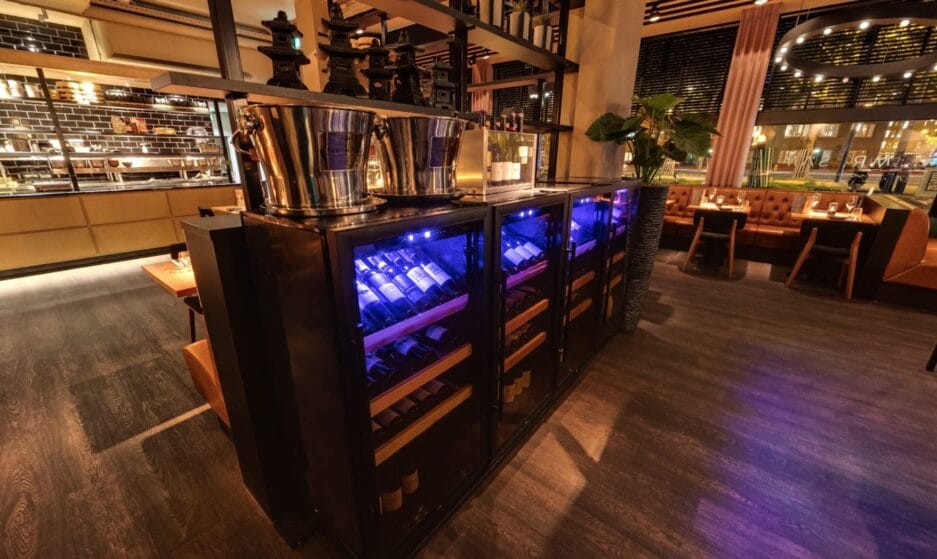 Interieur van een restaurant met wijnbewaarkasten met blauw licht, houten plateaus en wijnkoelers erboven op geplaatst.