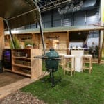 A modern outdoor kitchen at a fair, with a kitchen layout with wood paneling, a bar, stools and a green ceramic grill.