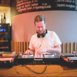 Ein DJ mischt Musik auf Plattenspielern in einer warm beleuchteten Barumgebung, umgeben von Schallplatten und einer Tafelkarte im Hintergrund.