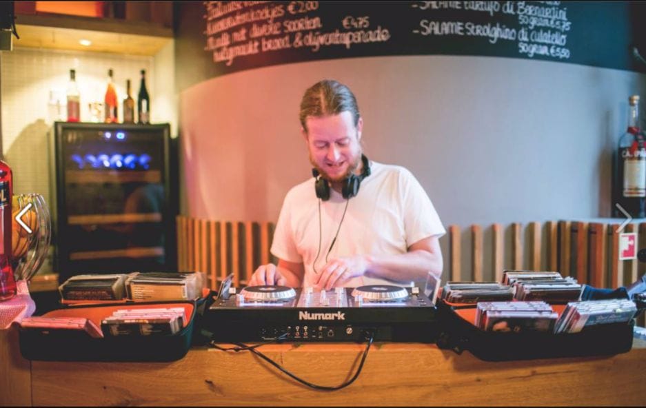 Een dj die muziek mixt op draaitafels in een warm verlichte baromgeving, omringd door vinylplaten en een schoolbordmenu op de achtergrond.