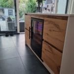A modern wine cooler with glass door and blue interior lighting, integrated into a wooden cabinet in a bright room with large windows.