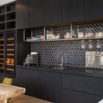 Cuisine moderne avec armoires noires, mur arrière en chevron, appareils électroménagers intégrés et table à manger en bois.
