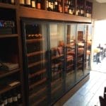 Ein Weinkühlschrank mit Glastüren neben mit Flaschen gefüllten Holzregalen in einer schwach beleuchteten Restaurantumgebung.