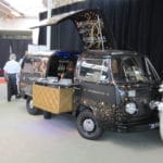 A vintage Volkswagen van converted into a mobile bar, called 'bubbles & co', during an indoor event with a server in white and black clothing nearby.
