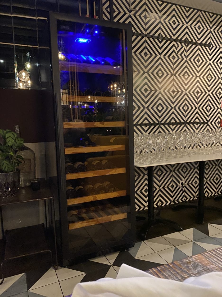 Interieur van een restaurant met een muur met decoratieve patronen en een glazen wijnkast die 's nachts een straat buiten weerspiegelt.