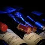 Een reeks wijnflessen met vintage etiketten, schuin tentoongesteld bij weinig licht, met de nadruk op rode lakzegels en reliëfdetails.