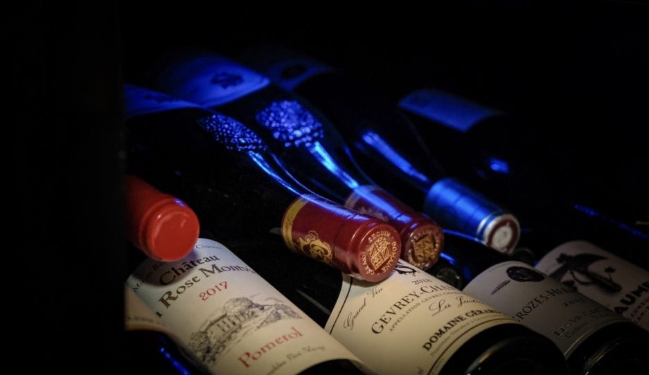 Een reeks wijnflessen met vintage etiketten, schuin tentoongesteld bij weinig licht, met de nadruk op rode lakzegels en reliëfdetails.