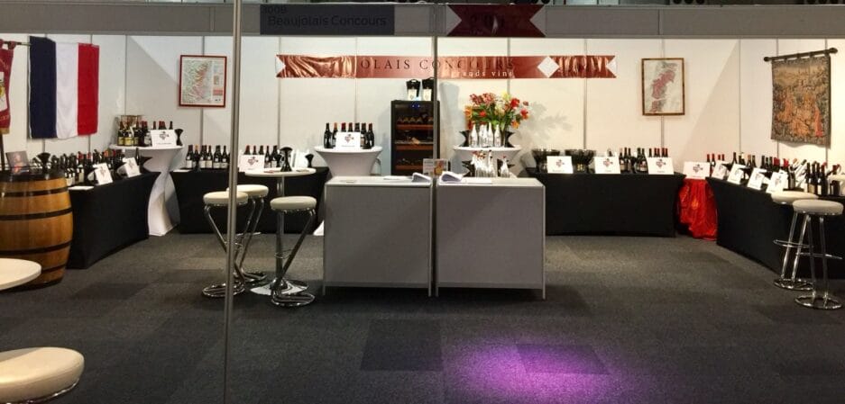 Een wijnproeverij op een beurs met flessen op tafels, krukken om op te zitten en spandoeken met de tekst 'beaujolais concours'.