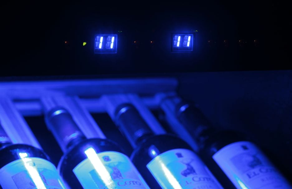 A wine rack illuminated by blue light, with several bottles, with digital clocks glowing on the dark background.