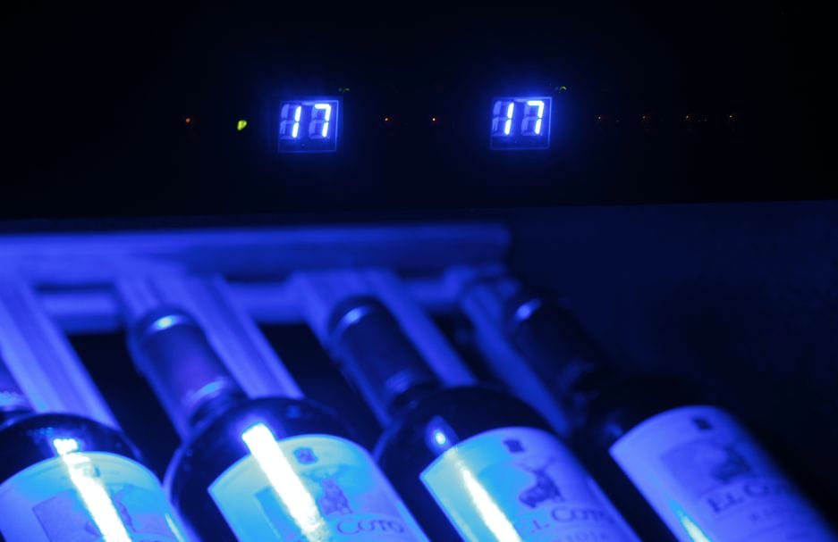 Digital clocks with "17:17" in blue lighting above wine bottles with blue lighting in a rack.