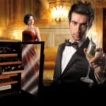 Ein Mann im Smoking hält ein Champagnerglas in der Hand, während eine Frau in einem roten Kleid neben einem Weinkühler in einem opulenten Raum sitzt.