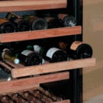 Een verscheidenheid aan wijnflessen horizontaal gestapeld op houten rekken in een koelkast met glazen front.