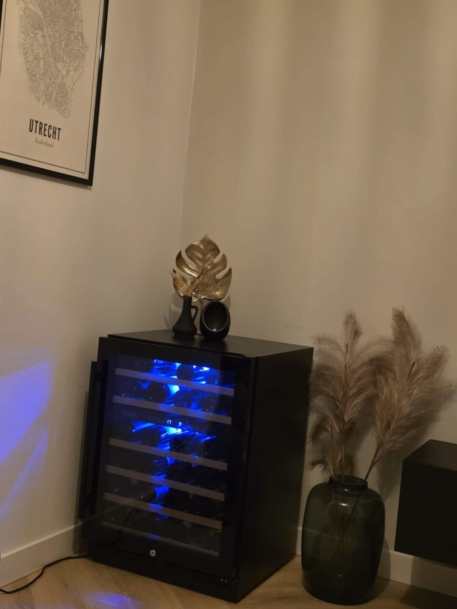 De wijnklimaatkast met mooie blauwe binnenverlichting staat in de hoek, naast een donkere vaas met pampagras en een decoratief bladgoud erop. Aan de muur hangt een ingelijste Utrechtse kaart.