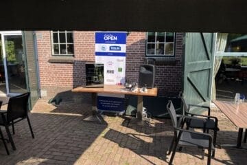 Buitenopstelling met een tafel, stoelen, speaker en promotionele banners onder een groene luifel. Bakstenen gebouw en open deur op de achtergrond.