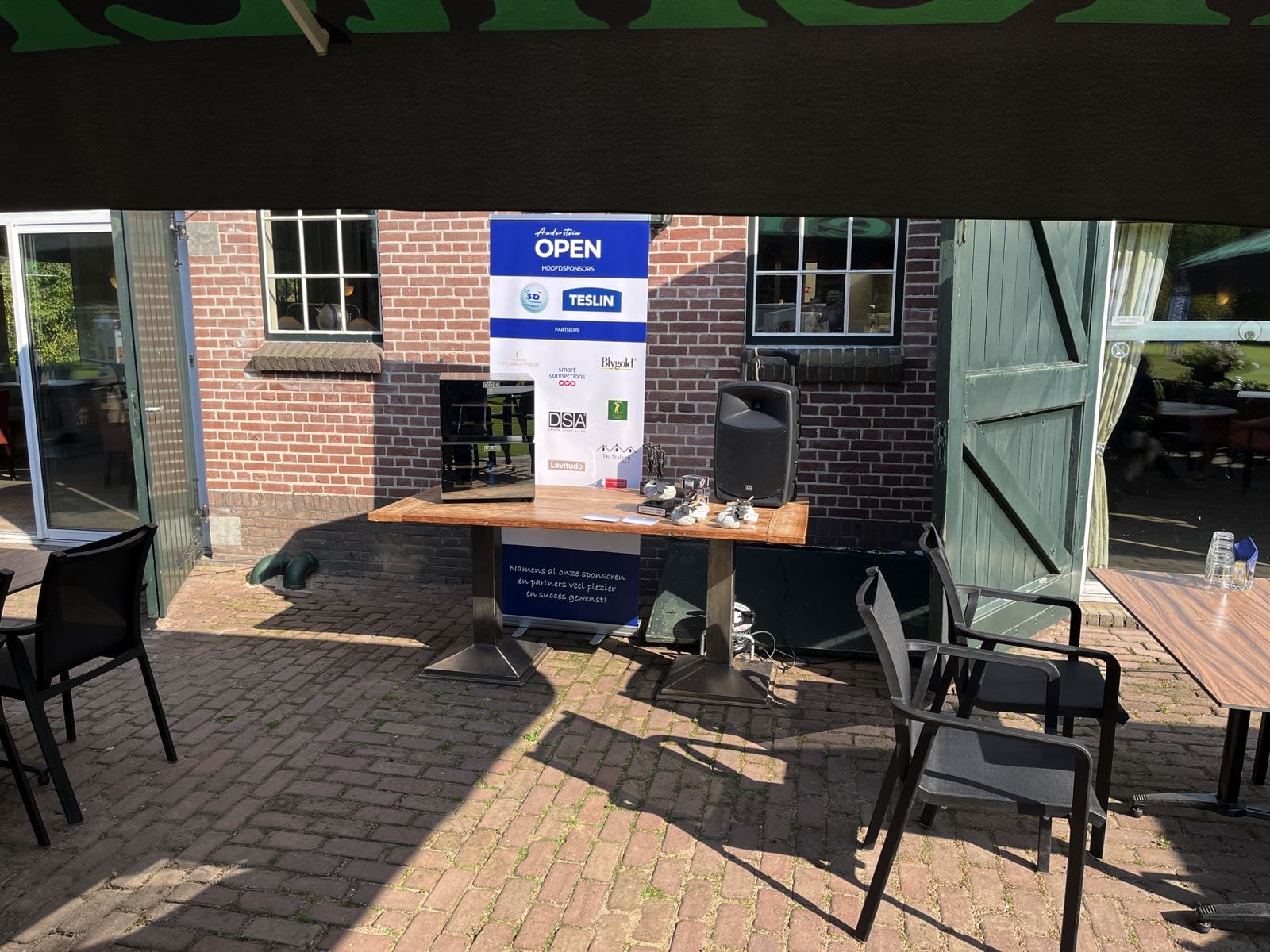 Outdoor setup with table, chairs, speaker and promotional banners under green canopy. Brick building and open door in the background.