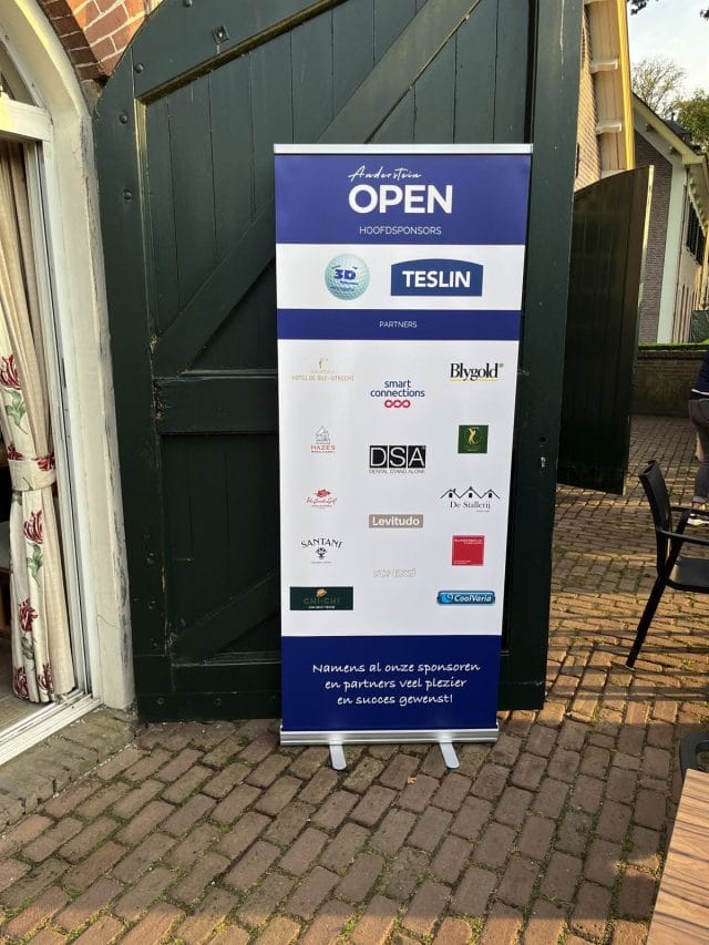 Banner met bovenaan "OPEN Hoofdsponsors", diverse sponsorlogo's en namen tegen een groene achtergrond van de deur, op een stenen pad.