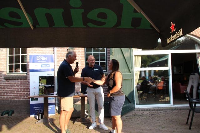 Drie mensen praten onder een tentluifel op een buitenevenement, met twee op de voorgrond en één met een microfoon. Een bakstenen gebouw en evenementenbanner zijn zichtbaar op de achtergrond.