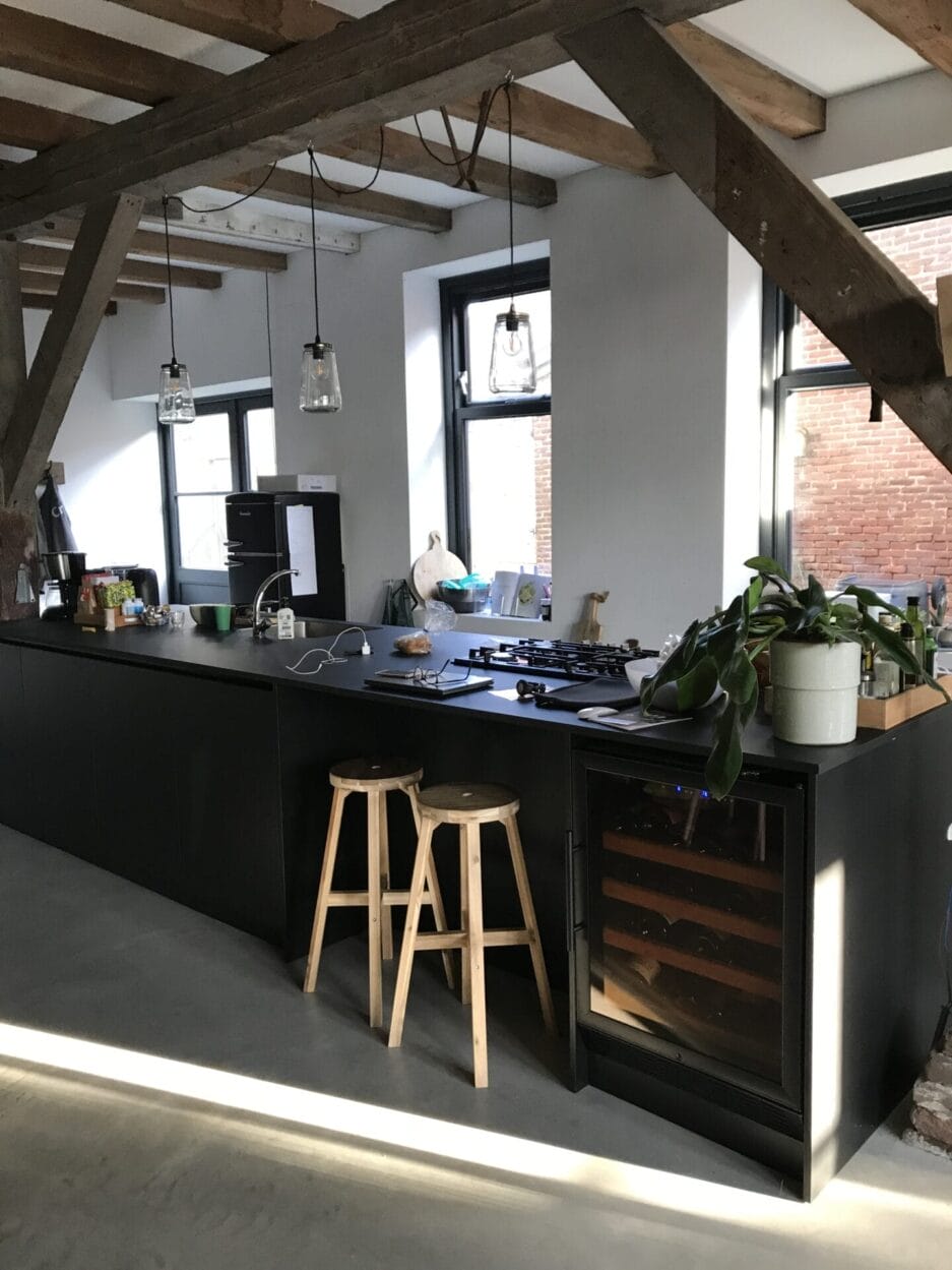 Een moderne keuken met een zwart aanrechtblad, barkrukken, zichtbare houten balken en hangende verlichting in een ruimte in loftstijl.