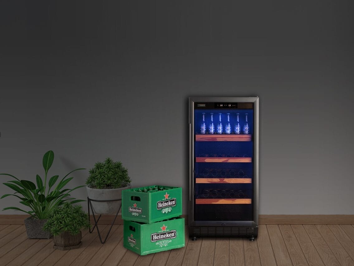 Een bier koelkast met glazen deur staat vol met flessen bier op de bovenste plank, naast twee groene Heineken-dozen. Potplanten staan ernaast op een houten vloer tegen een donkergrijze muur.