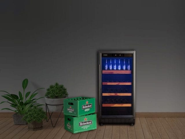 Een bier koelkast met glazen deur staat vol met flessen bier op de bovenste plank, naast twee groene Heineken-dozen. Potplanten staan ernaast op een houten vloer tegen een donkergrijze muur.