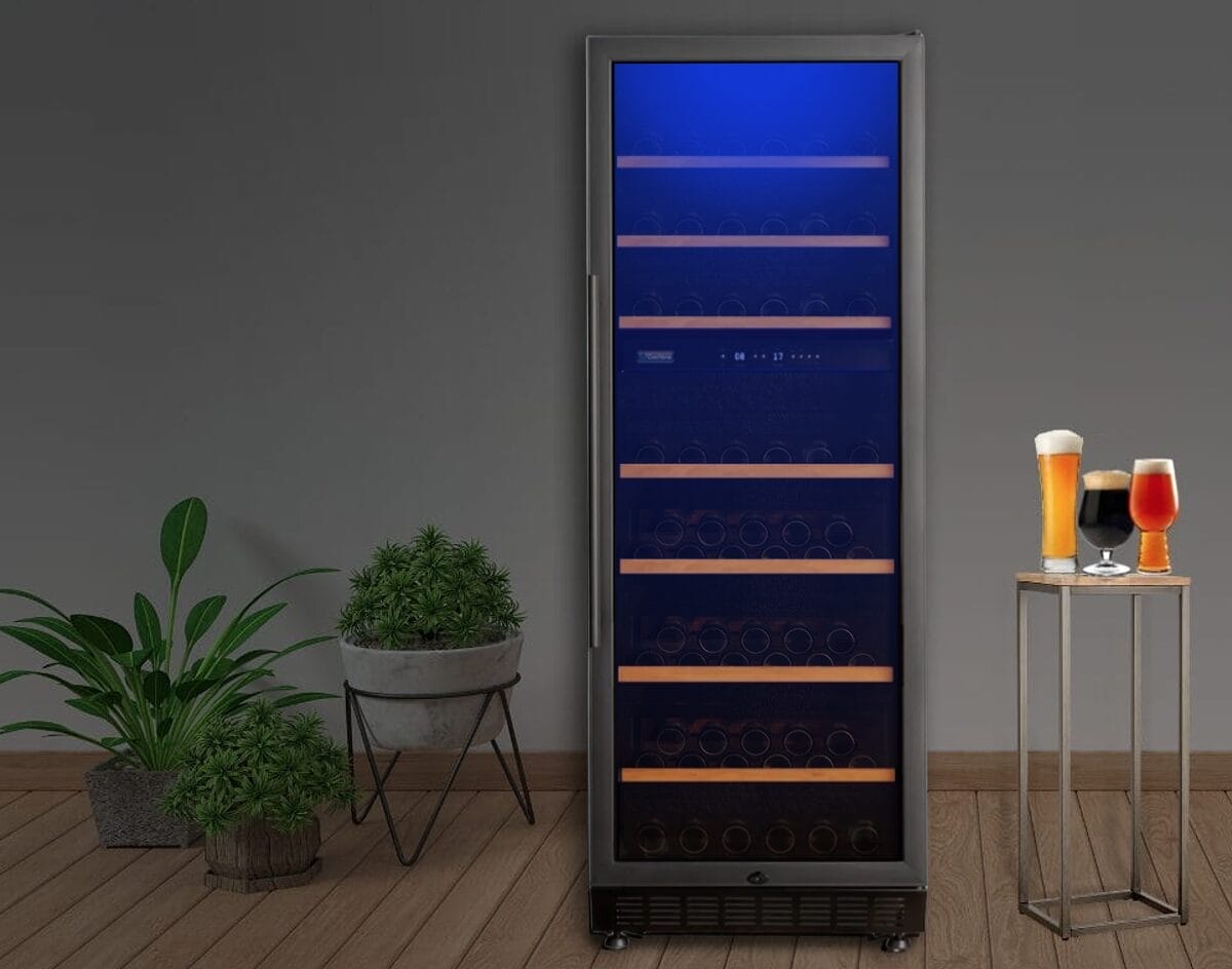 Grand refroidisseur à bière avec porte vitrée sur laquelle les bouteilles sont exposées sous une lumière bleue. Deux plantes en pot à gauche et deux verres de bière sur une petite table à droite contre un mur uni.