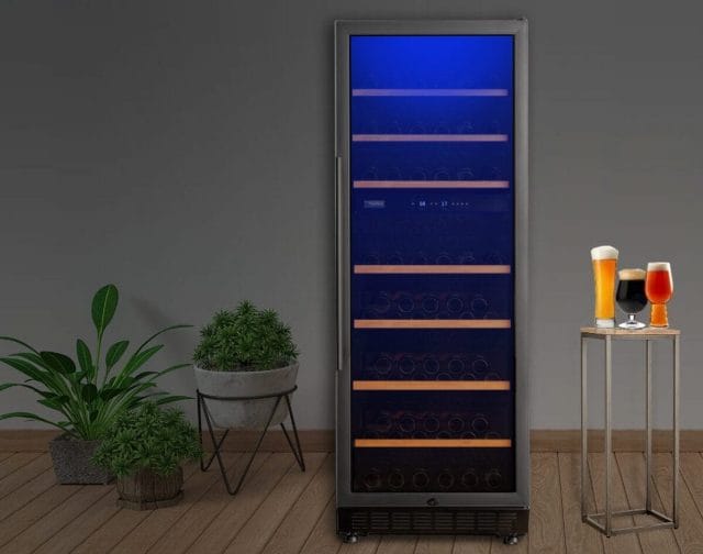 Tall beer cooler with glass door on which bottles stand under blue light. Two potted plants on the left and two glasses of beer on a small table on the right against a plain wall.