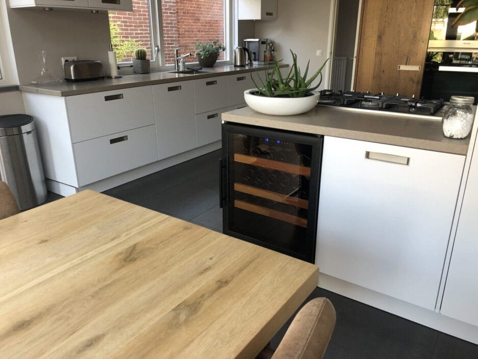 Moderne keuken met witte kasten, ingebouwde wijnkoelkast, houten tafel en een gasfornuis. Het decor omvat een schaal met groene appels op het aanrecht.