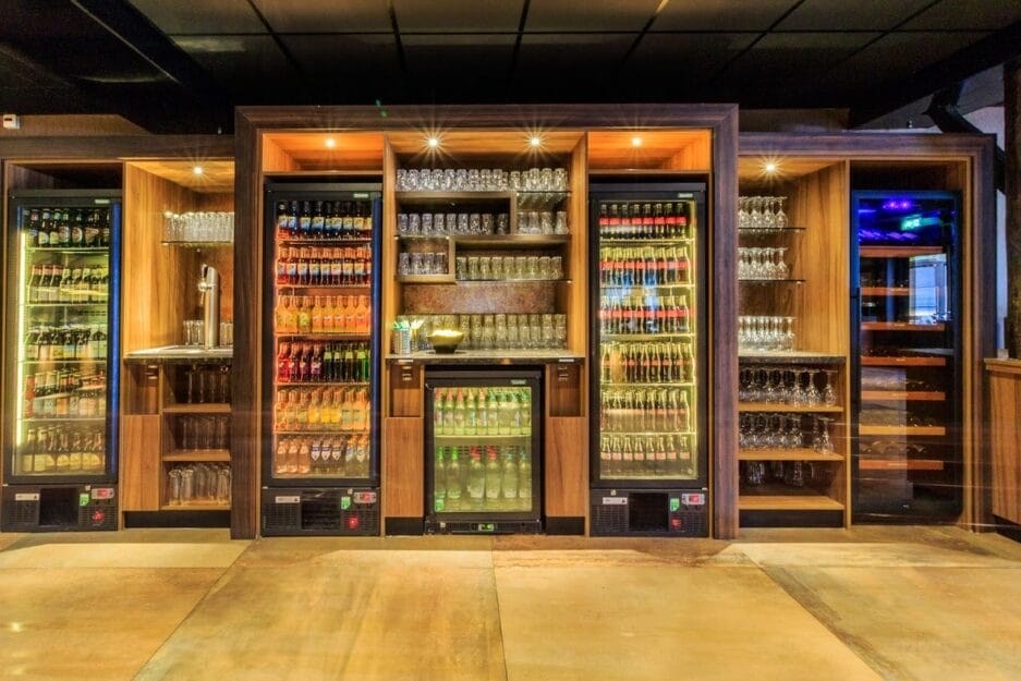 Modern barinterieur met meerdere koelkasten en een bierbewaarkast volledig gevuld met diverse flessen, geflankeerd door houten planken gevuld met glazen.