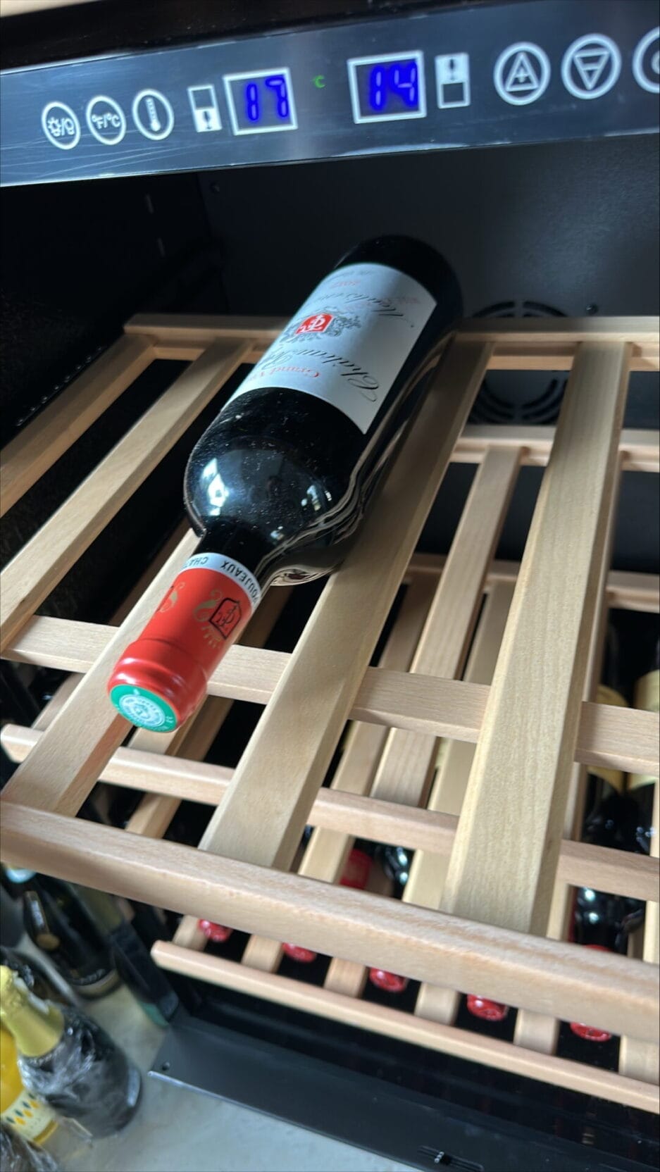 Een enkele fles rode wijn wordt horizontaal op een houten rek geplaatst in een wijnkoeler met digitale temperatuurdisplays (met nog een beschermfolie op het display) die 18°C en 14°C aangeven.