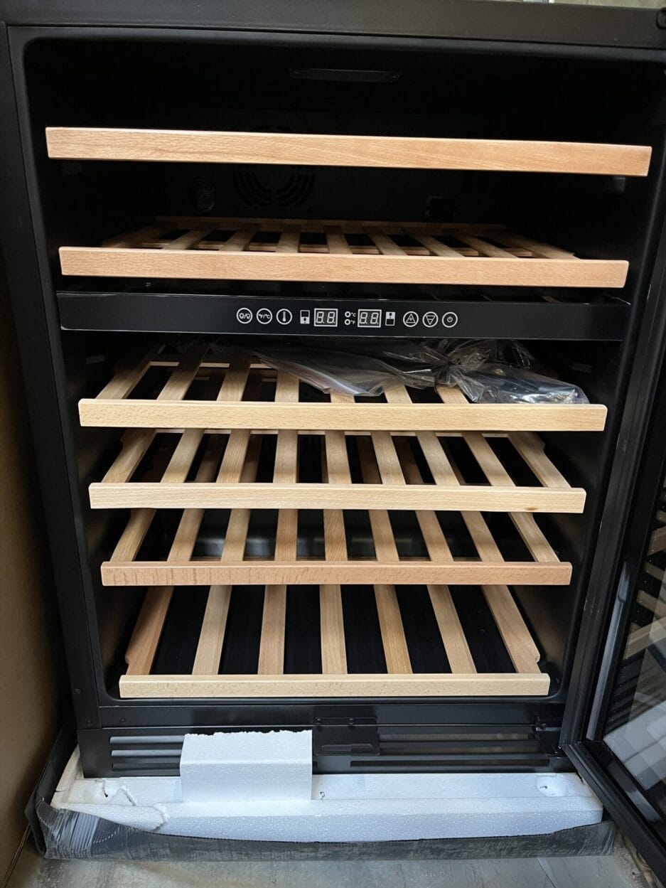 Een wijnklimaatkast met vijf houten uitschuifbare plateaus + éen opslagplateau, digitale bediening aan de binnenkant en de deur open, waardoor het lege interieur zichtbaar is, en in nog voorzien van transport verpakking.