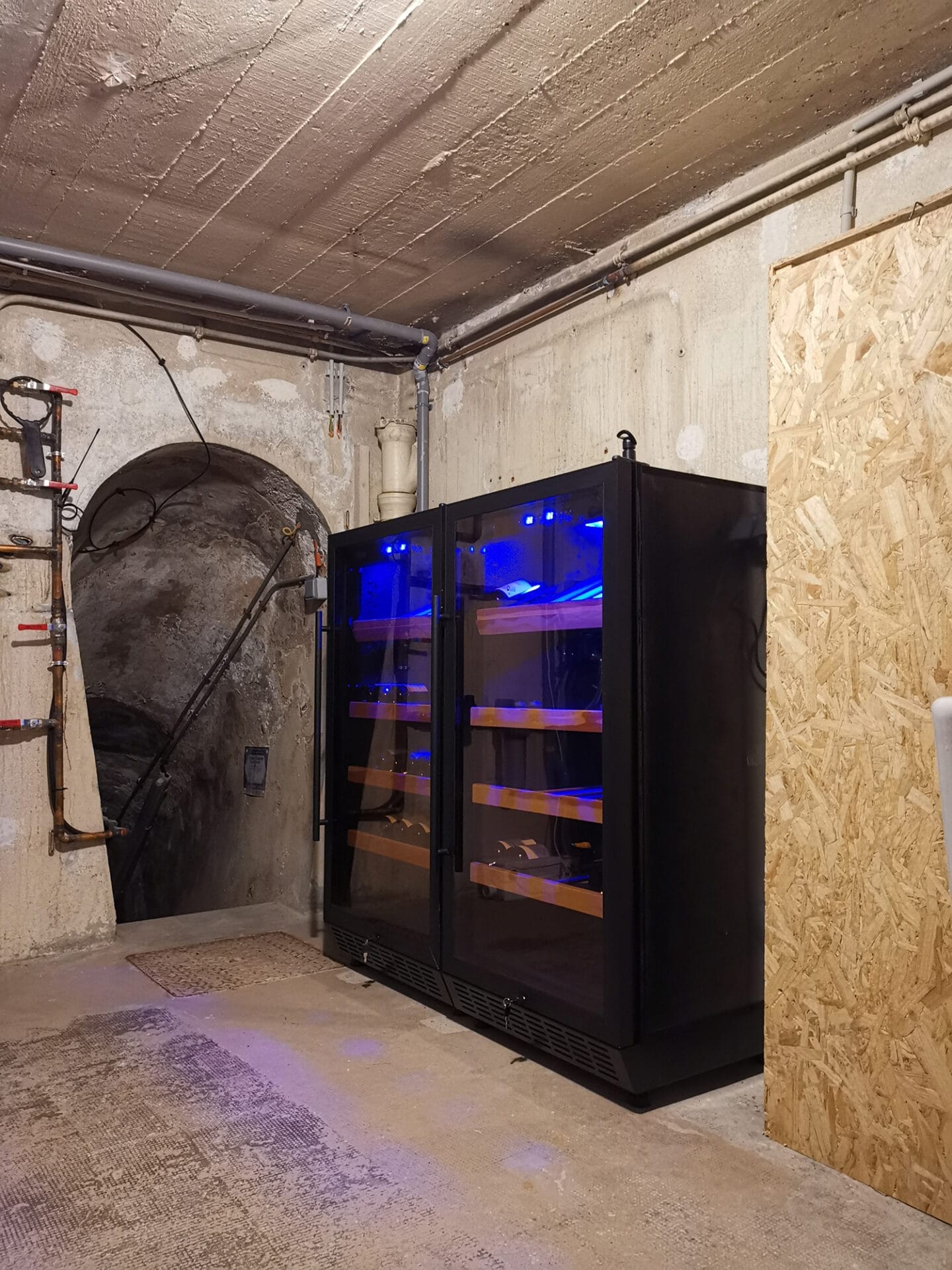 Een wijnkelder voorzien van twee wijnbewaarkasten, met blauwe verlichting in een verlichte, onafgewerkte kelder.