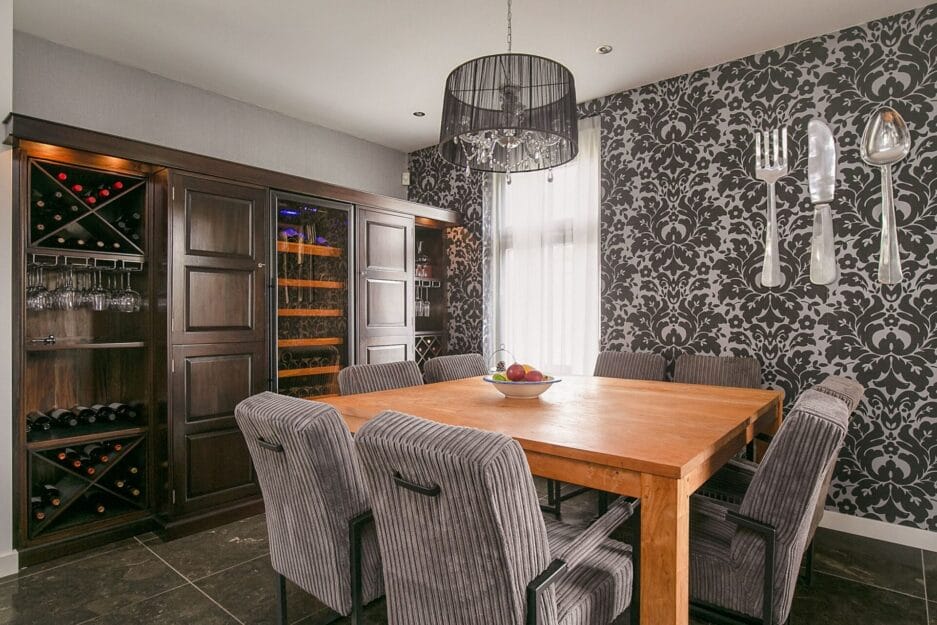 Een moderne eetkamer met een houten tafel, gestreepte stoelen, patroonbehang en een wijnkast. erboven hangt een kroonluchter.