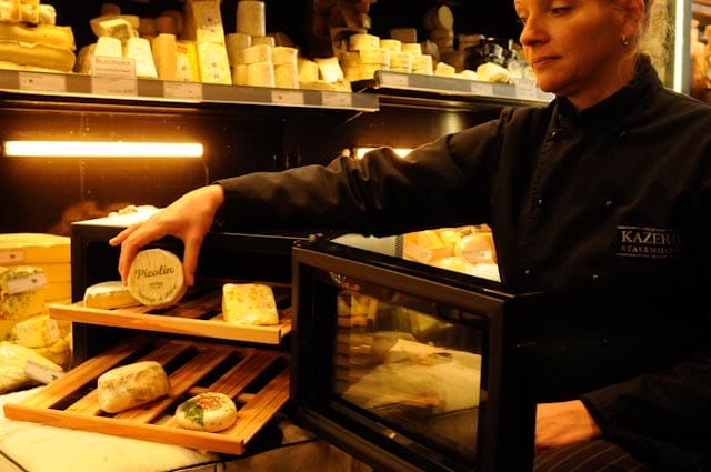 Un fromager en uniforme noir dispose divers fromages sur des étagères en bois dans un magasin faiblement éclairé à l'aide de l'armoire climatique à fromages (25 litres + deux planches à fromages).