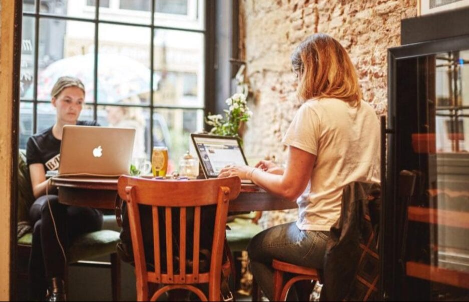 Twee mensen werken op laptops aan aparte tafels in een gezellig café met bakstenen muren, voorzien van een bierbewaarkast.