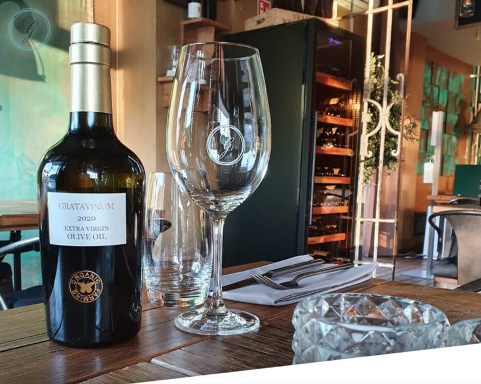 Een fles gratavinum extra vergine olijfolie en een leeg wijnglas op de tafel van een restaurant, met een wijnkoeler op de achtergrond.