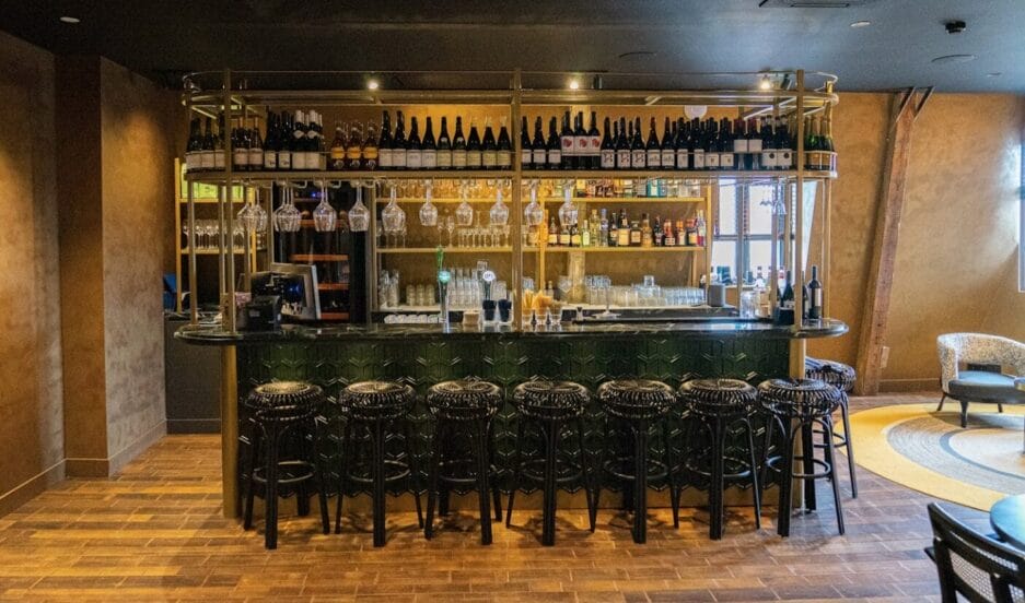 Een lege bar met krukken, vol met verschillende flessen alcohol en glazen, en een goed verlichte plank met extra flessen.