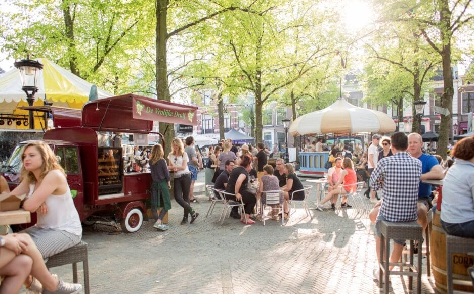 Buitenmarktscène met mensen die op een zonnige dag rond eetkraampjes onder bomen met elkaar praten. De "wijntruck" is voorzien van een wijn bewaarkast.