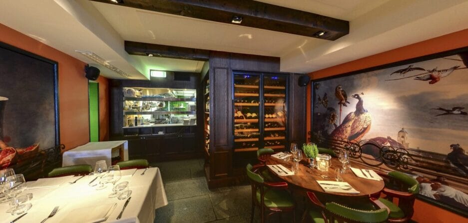 Interieur van een gezellig restaurant met een rustieke inrichting, een grote muurschildering van vogels, levendige oranje muren, zichtbare houten balken en een wijnkast.