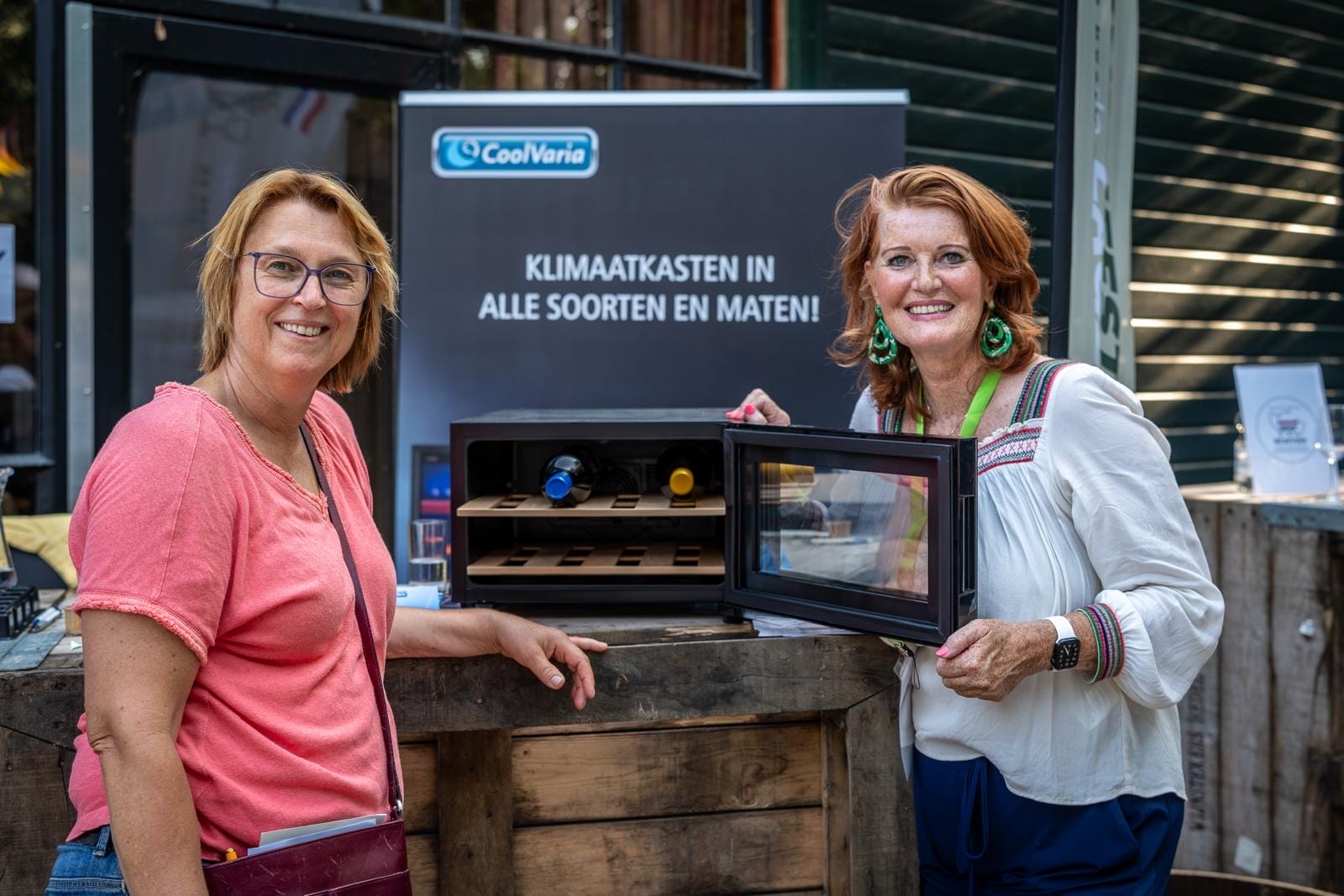 Twee vrouwen staan naast een open koelkastje met daarop flessen wijn, met een spandoek erachter.