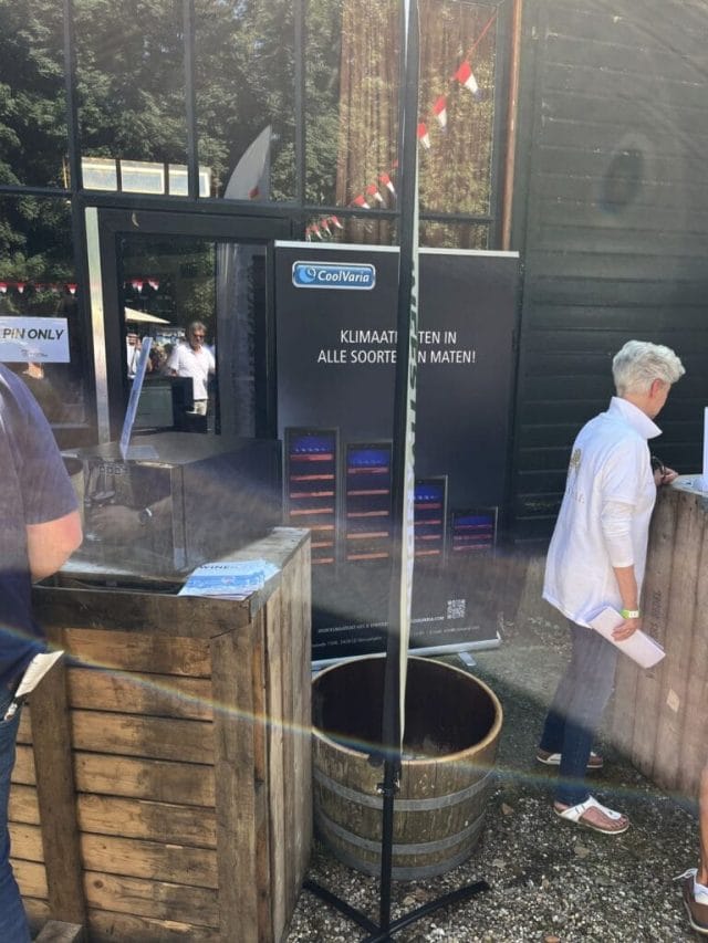 A person with short hair stands at a wooden table near a display titled "Klimaan." A barrel and wooden crates sit nearby, with trees and festive streamers in the background.