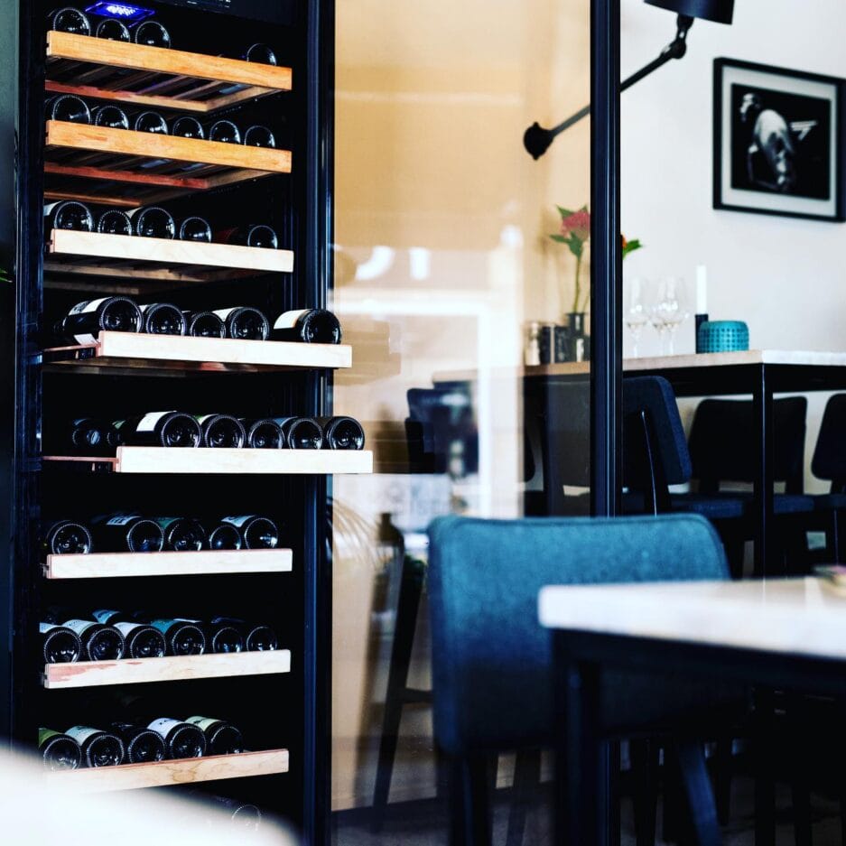 Interieur van een restaurant met een wijnrek met glazen voorkant gevuld met flessenwijn, zichtbaar door een deuropening; tafels en stoelen op de voorgrond.