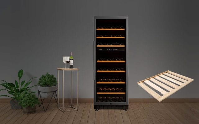 Grand réfrigérateur à vin rempli de bouteilles, posé sur un plancher en bois. Une petite table avec deux verres à vin et une bouteille se trouve à côté. Deux plantes en pot se trouvent à gauche. Un tiroir à vin vide flotte.