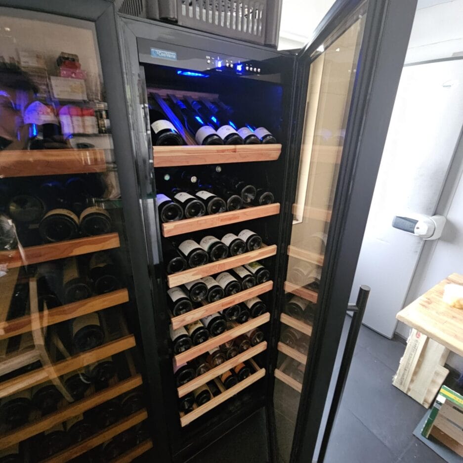 Open wijnkoelkast gevuld met rijen horizontaal opgeslagen wijnflessen, zichtbaar door glazen deuren in een kamer.