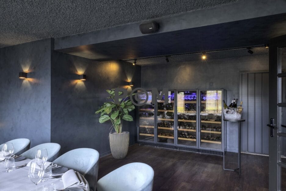 Een rij wijnbewaarkasten (in een restaurant) met glazen fronten gevuld met flessen staat tegen een donkere muur naast een potplant en een tafeltje met een ijsemmer met wijnflessen.