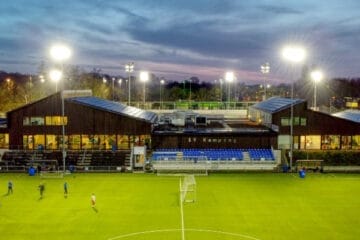 A brightly lit soccer field at sunset with players warming up. A modern sports facility with seating is in the background.