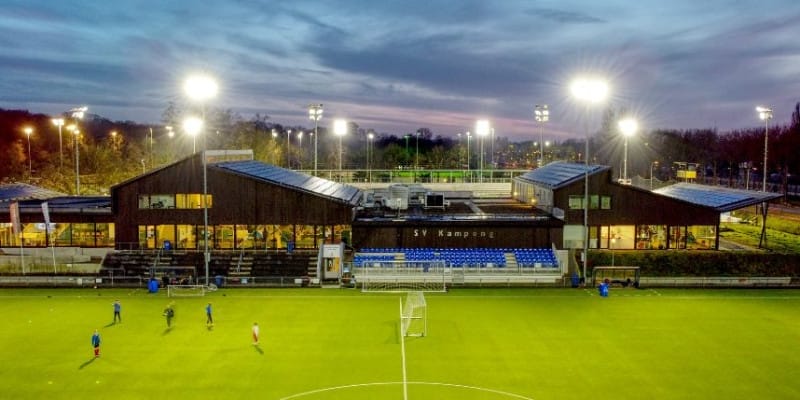 A brightly lit soccer field at sunset with players warming up. A modern sports facility with seating is in the background.